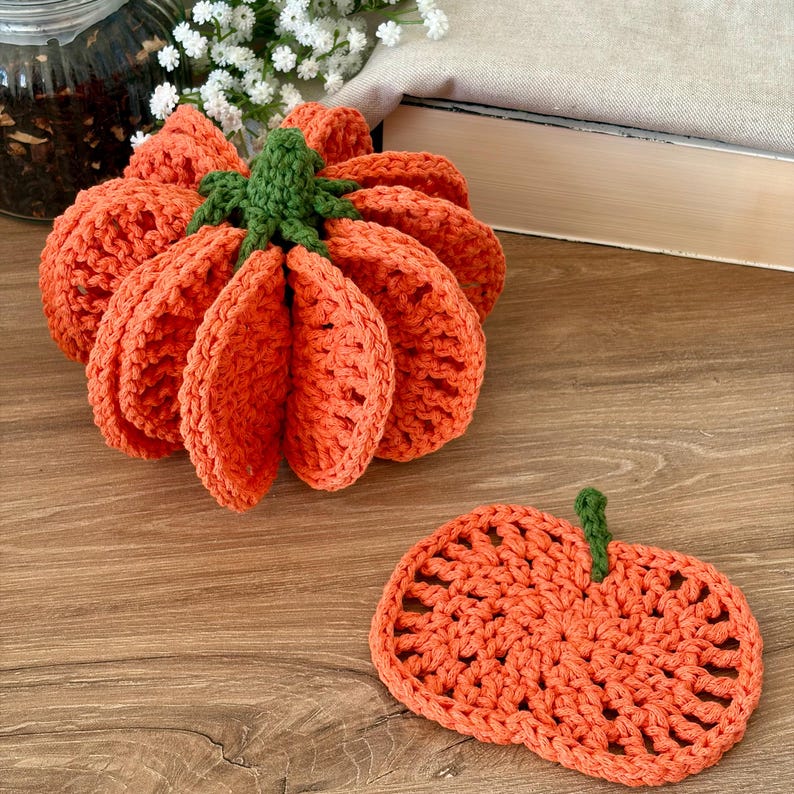 May include: A three-dimensional crocheted orange pumpkin with a green stem and a matching flat, circular crocheted orange pumpkin coaster with a small green stem, both featuring an open, textured stitch pattern.