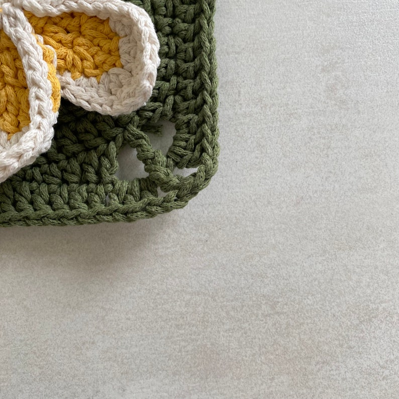 May include: A green crocheted square with a white and yellow crocheted flower on top.