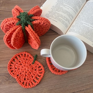 May include: A set of orange crochet pumpkin-shaped coasters with green stems. One coaster is stacked in a folded bundle, while another lies flat beneath a white ceramic mug. The coasters feature a circular, open-weave pattern with a textured finish.