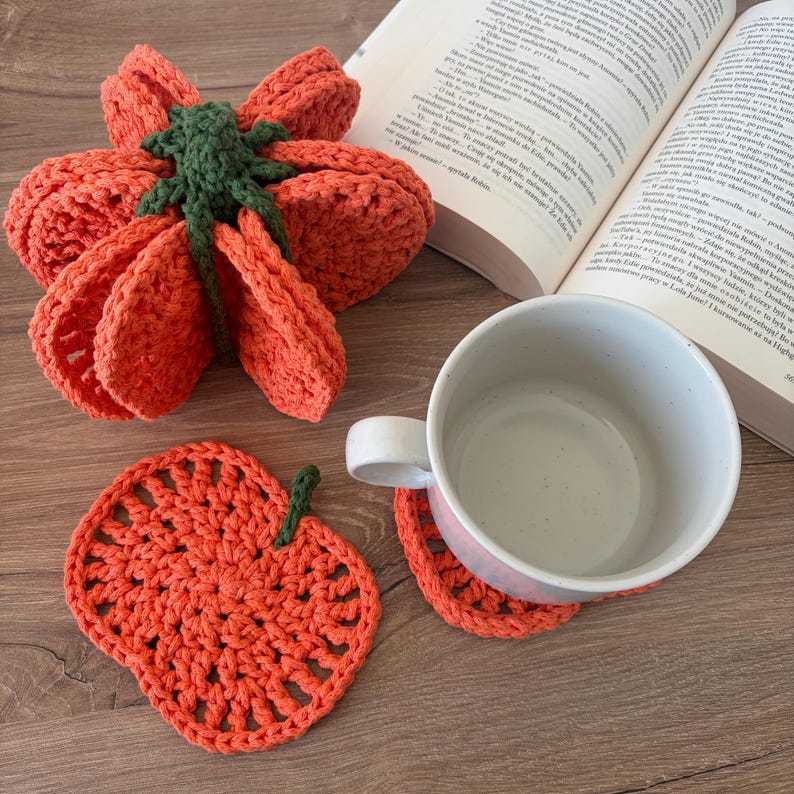 May include: A set of orange crochet pumpkin-shaped coasters with green stems. One coaster is stacked in a folded bundle, while another lies flat beneath a white ceramic mug. The coasters feature a circular, open-weave pattern with a textured finish.