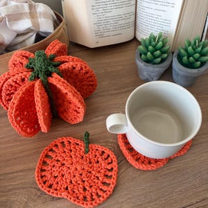 May include: A set of orange pumpkin-shaped crochet coasters. One coaster is stacked in a bundle with a green stem, while another lies flat. A white ceramic mug sits on a third coaster. The coasters feature a textured, open-stitch pattern.