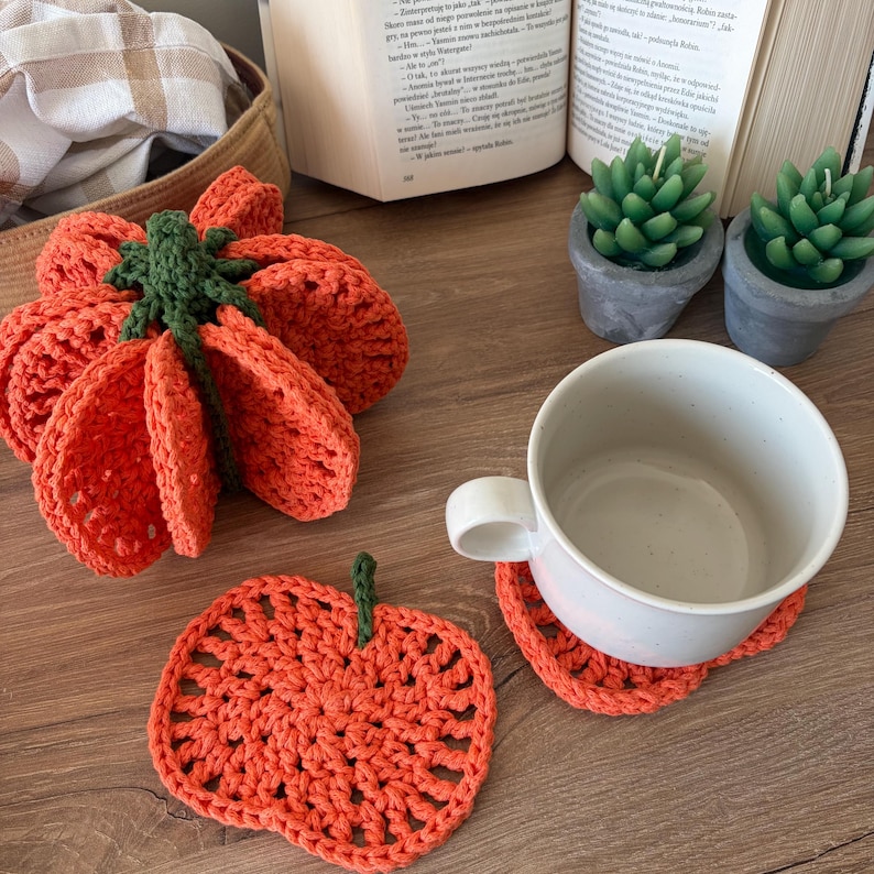 May include: A set of orange pumpkin-shaped crochet coasters. One coaster is stacked in a bundle with a green stem, while another lies flat. A white ceramic mug sits on a third coaster. The coasters feature a textured, open-stitch pattern.