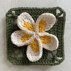 May include: A crocheted flower with five petals in shades of white and yellow, set against a green crocheted square background.