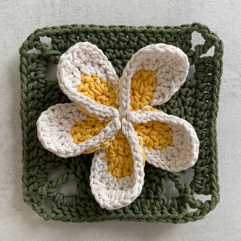 May include: A crocheted flower with five petals in shades of white and yellow, set against a green crocheted square background.