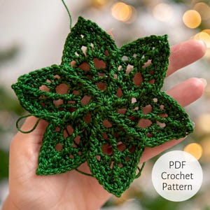 以下が含まれることがあります： 手作りの緑色の鉤針編みの星、五芒星のデザイン。星は手で持たれており、複雑な透かし模様を示しています。画像には「PDF Crochet Pattern」というテキストが入った白い円が含まれています。