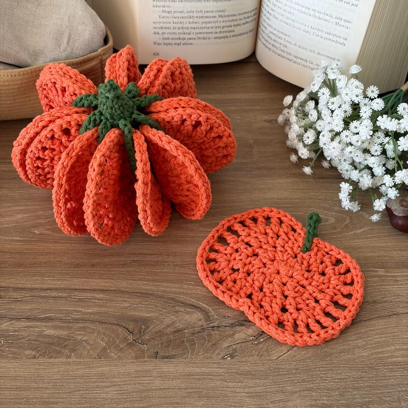 May include: A three-dimensional crocheted orange pumpkin with a dark green stem and a matching flat, circular crocheted orange pumpkin coaster with a small green stem, both featuring an open, airy stitch pattern throughout the design.