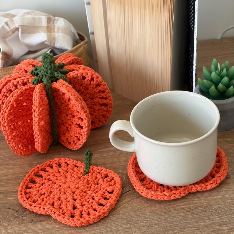 May include: A three-dimensional crocheted orange pumpkin with a green stem, alongside a flat, pumpkin-shaped orange crocheted coaster with a green stem, and a white ceramic mug resting on a second identical orange pumpkin-shaped coaster.