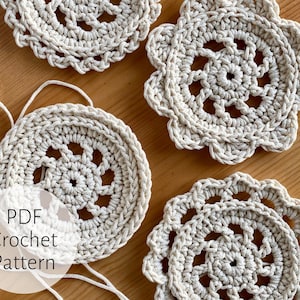 May include: Four crocheted coasters in a cream color with a delicate floral pattern. The coasters are arranged on a wooden surface. The text "PDF Crochet Pattern" is visible in the image.