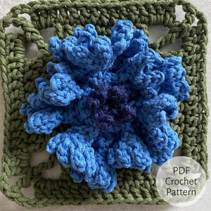 Könnte beinhalten: Handgefertigte Häkelblume in Blau- und Marineblautönen, zentriert auf einem grünen Quadrat. Die Blume hat detaillierte Blütenblätter und ein strukturiertes Aussehen. Das Quadrat hat ein Spitzenmuster. Der Text auf dem Bild lautet "PDF Crochet Pattern".