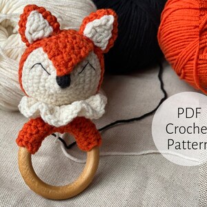 May include: Crocheted fox rattle toy with a wooden ring. The toy is orange, white, and black. The toy is surrounded by balls of yarn in white, black, and orange.