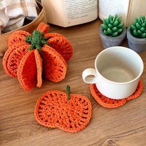 May include: A crocheted orange pumpkin with a green stem, a flat orange pumpkin-shaped coaster, and a white ceramic mug resting on a second orange pumpkin coaster. The items are crafted from yarn with visible stitching and textured patterns.
