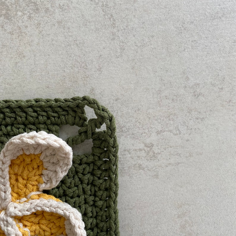 May include: A crocheted green and white flower with yellow accents. The flower is part of a larger crocheted piece.