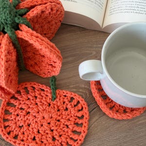 May include: A set of orange crochet pumpkin-shaped coasters with green stems. One coaster is flat on the surface, while another is stacked underneath a white ceramic mug. The coasters feature a circular, open-weave stitch pattern in bright orange yarn.