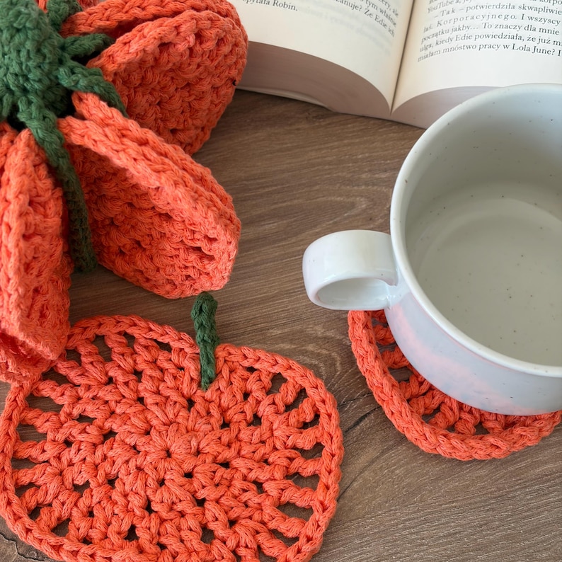 May include: A set of orange crochet pumpkin-shaped coasters with green stems. One coaster is flat on the surface, while another is stacked underneath a white ceramic mug. The coasters feature a circular, open-weave stitch pattern in bright orange yarn.