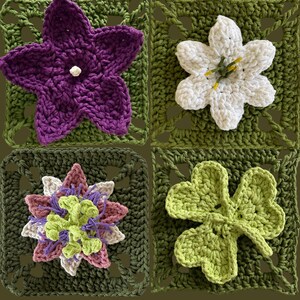 May include: Four crocheted squares with floral designs. The top left square features a purple flower with a white center. The top right square has a white flower with yellow stamens. The bottom left square has a multi-colored flower. The bottom right square has a green shamrock.
