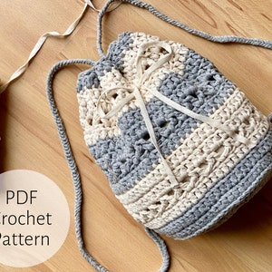 May include: A cream and grey crochet drawstring bag with a striped design. The bag has a cream-coloured drawstring closure. The image includes the text "PDF Crochet Pattern". The bag is displayed on a wooden surface.