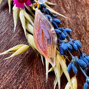 Può includere: Un ciondolo in resina trasparente con una cauzione color oro, caratterizzato da un design a strati con sfumature rosse e viola. Il ciondolo è adornato con un piccolo fiore rosa, fiori blu e fogliame verde e giallo, su uno sfondo di legno scuro.
