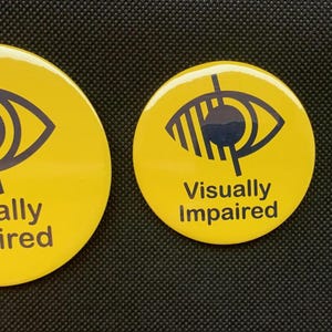 以下が含まれることがあります： 黒い目のシンボルと「Visually Impaired」というテキストが書かれた、黄色い丸いボタンが3つあります。ボタンはサイズが異なります。