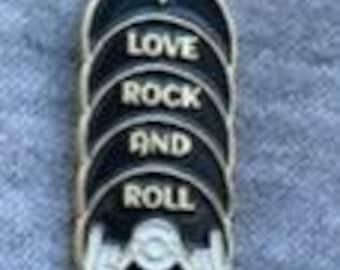 May include: An oval-shaped metal pin with a black background and silver-toned raised lettering that reads I LOVE ROCK AND ROLL. The bottom of the pin features a silver-toned graphic of a hand making the rock and roll sign with fingers extended.