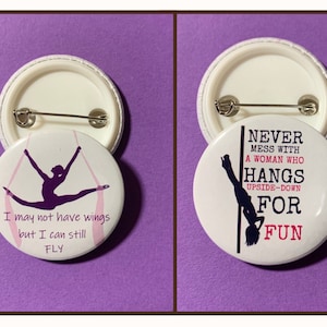 May include: Two round white button pins with a black silhouette of a person hanging upside down. The pin on the left says "I may not have wings but I can still FLY". The pin on the right says "NEVER MESS WITH A WOMAN WHO HANGS UPSIDE-DOWN FOR FUN".