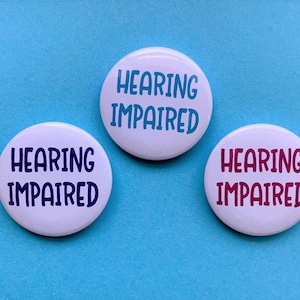 Op de afbeelding: Drie witte ronde knoppen met de tekst "Hearing Impaired" in blauwe, paarse en roze letters.