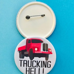 Puede incluir: Dos insignias redondas. Una insignia es blanca con un imperdible en la parte posterior. La otra insignia presenta un gráfico de camión rojo y las palabras "TRUCKING HELL!" en negro.
