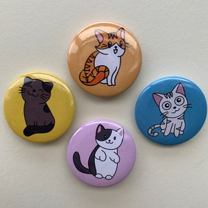 May include: Four round pinback buttons featuring different cartoon cats. One button has a brown cat with white eyes, one has a black and white cat with pink ears, one has a brown and orange cat with white eyes, and one has a white cat with black and orange markings.