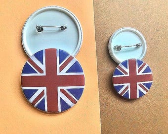 May include: Two round pinback buttons featuring the Union Jack flag design in red, white, and blue. The buttons are on a light orange and brown background. The backs of the buttons are white.