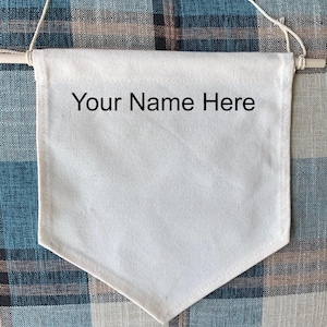 May include: A plain, cream-coloured banner with the text "Your Name Here" in black. The banner is rectangular with a pointed base and is hung from a wooden dowel with a string. The background features a blue and grey plaid pattern.