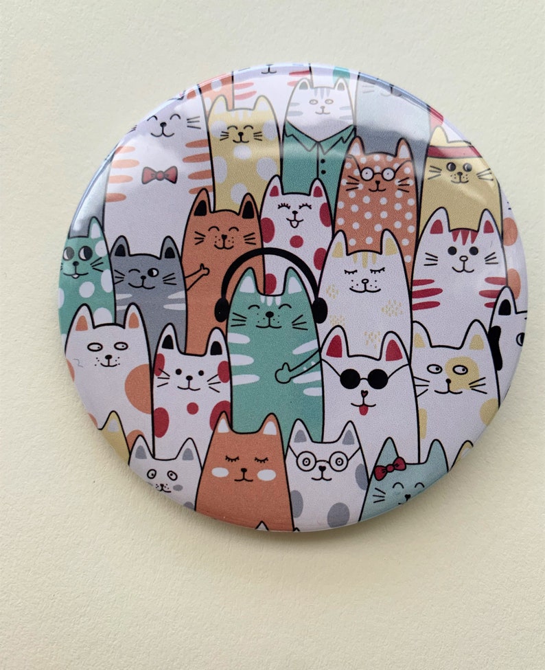 K&ouml;nnte beinhalten: Ein runder Spiegel mit einem farbenfrohen Design von Katzen, die verschiedene Accessoires tragen. Die Katzen sind alle in verschiedenen Farben und haben unterschiedliche Gesichtsausdr&uuml;cke.