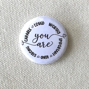 May include: A white badge with the words "you are" in a cursive font. The text is surrounded by the words "capable, loved, worthy, important, kind, strong" with heart symbols.