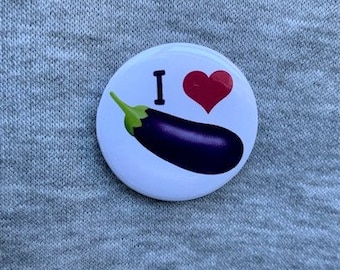 Könnte beinhalten: Ein weißer Knopf mit dem Text "Ich liebe" und einem roten Herzen über einer lila Aubergine.