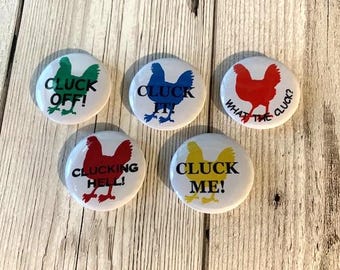 Könnte beinhalten: Fünf runde weiße Knöpfe mit einer schwarzen Hühnersilhouette und einem humorvollen Hühner-Slogan. Die Knöpfe sind grün, blau, rot, gelb und rot. Die Slogans sind "Cluck Off!" "Cluck It!" "What the Cluck?" "Cluck Me!" und "Cluckin' Hell!"