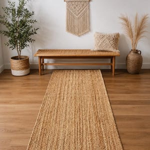 May include: A rectangular runner rug made of natural woven jute fibers with a textured, braided appearance. The rug features a consistent tan and light brown color palette, lying flat on a smooth wooden floor surface in a long, narrow shape.