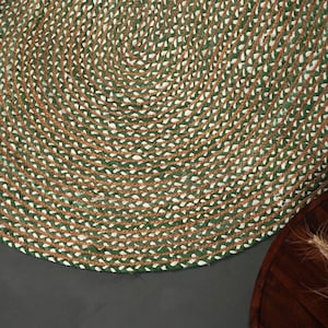 Può includere: Primo piano di un tappeto in juta intrecciata verde, marrone e bianco. Il tappeto ha una forma circolare con un motivo a spirale. Il tappeto è realizzato con fibre naturali e ha un aspetto strutturato. Una ciotola di legno con spighe di grano è sullo sfondo.