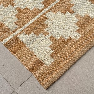 May include: A woven area rug featuring a natural tan jute texture with a repeating geometric pattern in off-white. The design consists of stepped, diamond-like shapes arranged in rows across the surface, showcasing a coarse, fibrous material construction.