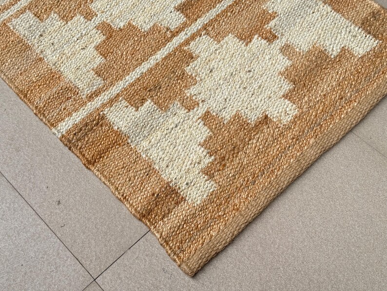 May include: A woven area rug featuring a natural tan jute texture with a repeating geometric pattern in off-white. The design consists of stepped, diamond-like shapes arranged in rows across the surface, showcasing a coarse, fibrous material construction.