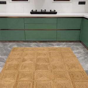 May include: A rectangular, woven jute rug with a geometric square pattern. The rug is a warm tan color and is placed on a gray tiled floor in a kitchen setting. The kitchen features green cabinets and a white countertop.