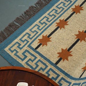 Könnte beinhalten: Ein gewebter Teppich mit blauem Rand mit einem geometrischen Muster. Der Teppich hat einen beigefarbenen Hintergrund mit orangefarbenen Sternenmustern und schwarzen diagonalen Streifen. Der Teppich hat einen Fransenrand.