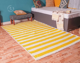 May include: A yellow and white striped rug with a white border. The rug is on a wooden floor in front of a gray couch with white and yellow pillows.