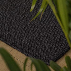 May include: A dark grey rectangular rug with a textured, woven pattern. The rug is placed on a light-coloured wooden floor, with green foliage in the foreground. The rug's edges are neatly finished.