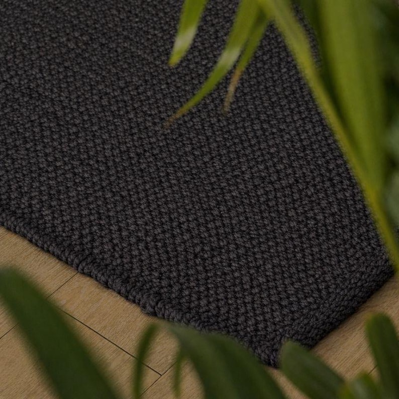 May include: A dark grey rectangular rug with a textured, woven pattern. The rug is placed on a light-coloured wooden floor, with green foliage in the foreground. The rug's edges are neatly finished.