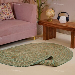 Puede incluir: Una alfombra redonda de yute trenzado con borde verde se encuentra sobre un suelo claro. La alfombra est&aacute; parcialmente desenrollada, revelando su dise&ntilde;o en espiral. Un sof&aacute; rosa y una mesa de centro de madera est&aacute;n en el fondo.