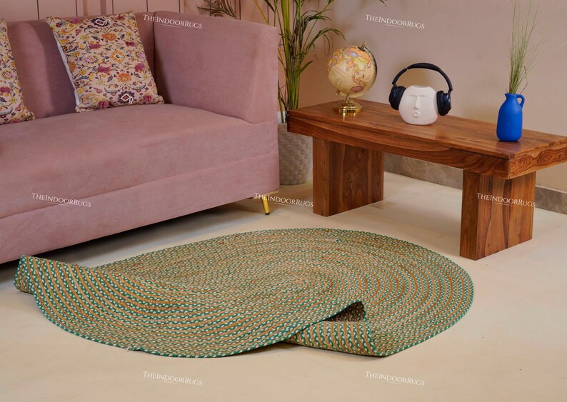Puede incluir: Una alfombra redonda de yute trenzado con borde verde se encuentra sobre un suelo claro. La alfombra est&aacute; parcialmente desenrollada, revelando su dise&ntilde;o en espiral. Un sof&aacute; rosa y una mesa de centro de madera est&aacute;n en el fondo.