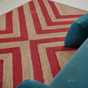 May include: A geometric jute rug with a red and natural color block design. The rug is placed next to a teal blue sofa. The rug's pattern is a series of red stripes that form a square pattern.