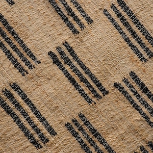 May include: Close-up of a tan textile with a repeating pattern of diagonal black stripes. The fabric has a textured, woven appearance, with visible fibers and a neutral color palette.
