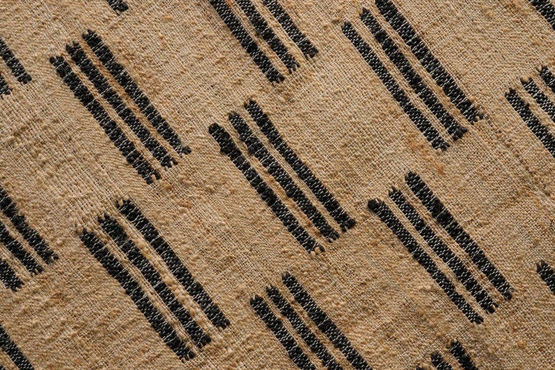 May include: Close-up of a tan textile with a repeating pattern of diagonal black stripes. The fabric has a textured, woven appearance, with visible fibers and a neutral color palette.