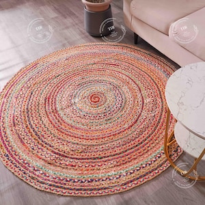 K&ouml;nnte beinhalten: Ein runder, mehrfarbiger geflochtener Jute-Teppich mit Spiralmuster. Der Teppich hat Farbt&ouml;ne von Orange, Rosa, Rot und Gr&uuml;n. Der Teppich liegt auf einem Holzboden, im Hintergrund sind ein Sofa und ein Beistelltisch zu sehen.