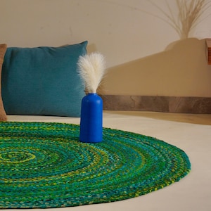 Può includere: Un tappeto rotondo intrecciato caratterizzato da un mix vivace di fibre verdi, verde acqua e gialle disposte in un motivo circolare. Il tappeto è accompagnato da un vaso cilindrico blu tinta unita contenente erbe decorative piumose bianche.
