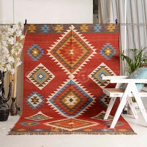 May include: A red rectangular rug with a geometric diamond pattern in shades of yellow, blue, and brown. The rug has a fringed edge and is displayed against a white background. The rug is a decorative floor covering.